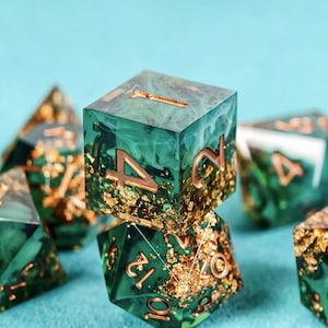 May include: A set of polyhedral dice made from translucent emerald green resin with swirling white smoke patterns and embedded gold leaf flakes. The numbers are engraved in a metallic copper finish, featuring a d20, d12, d10, d8, d6, and d4.
