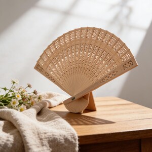 Puede incluir: Abanico plegable de madera clara con intrincados diseños calados, exhibido en un pequeño soporte de madera. El abanico está abierto, revelando su delicado patrón. Está sobre una mesa de madera con una tela beige y flores.