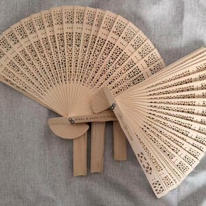 Puede incluir: Varios abanicos de madera clara abiertos, con intrincados diseños calados. Los abanicos están desplegados, mostrando los patrones detallados. Algunos abanicos tienen el texto "Aiden & Katie 7.16.2023" y "Anna & Ryan 10.15.2023".