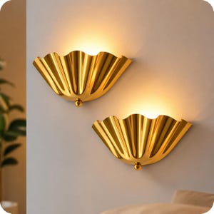 Puede incluir: Dos apliques de pared dorados con un diseño festoneado en forma de abanico. Cada aplique tiene un pequeño acento redondo en la parte inferior. Los apliques están montados en una pared gris claro, con una luz cálida que emana de ellos.