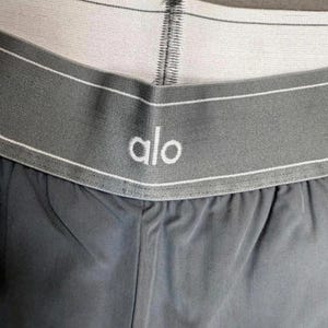 Puede incluir: Primer plano de unos pantalones cortos deportivos grises con una cintura blanca. La cintura presenta el nombre de la marca "alo" en letras blancas. Los pantalones cortos est&aacute;n hechos de una tela suave, posiblemente que absorbe la humedad.