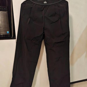 Puede incluir: Pantalones deportivos negros con dise&ntilde;o de pierna ancha. La cintura muestra el nombre de la marca "alo" en letras blancas. Los pantalones est&aacute;n colgados de una percha.