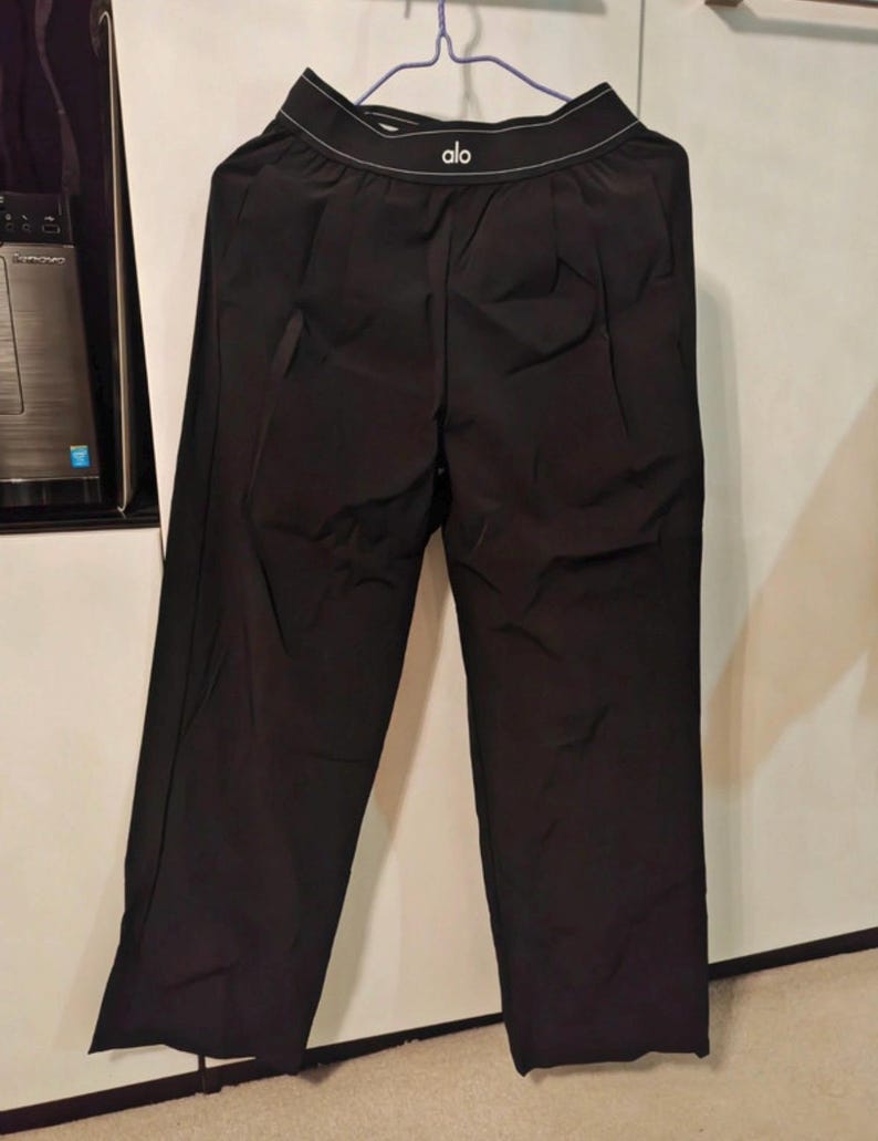 Puede incluir: Pantalones deportivos negros con dise&ntilde;o de pierna ancha. La cintura muestra el nombre de la marca "alo" en letras blancas. Los pantalones est&aacute;n colgados de una percha.