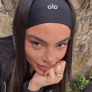 May include: Black headband with the white letters "olo" across the front. The headband is worn on the forehead. The person in the image is wearing a black jacket and a gold ring.
