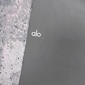 Puede incluir: Una esterilla de yoga gris oscuro con el texto blanco "alo" en la esquina superior izquierda. La esterilla es de color liso y parece estar hecha de un material liso. El fondo es una superficie texturizada de color gris claro.