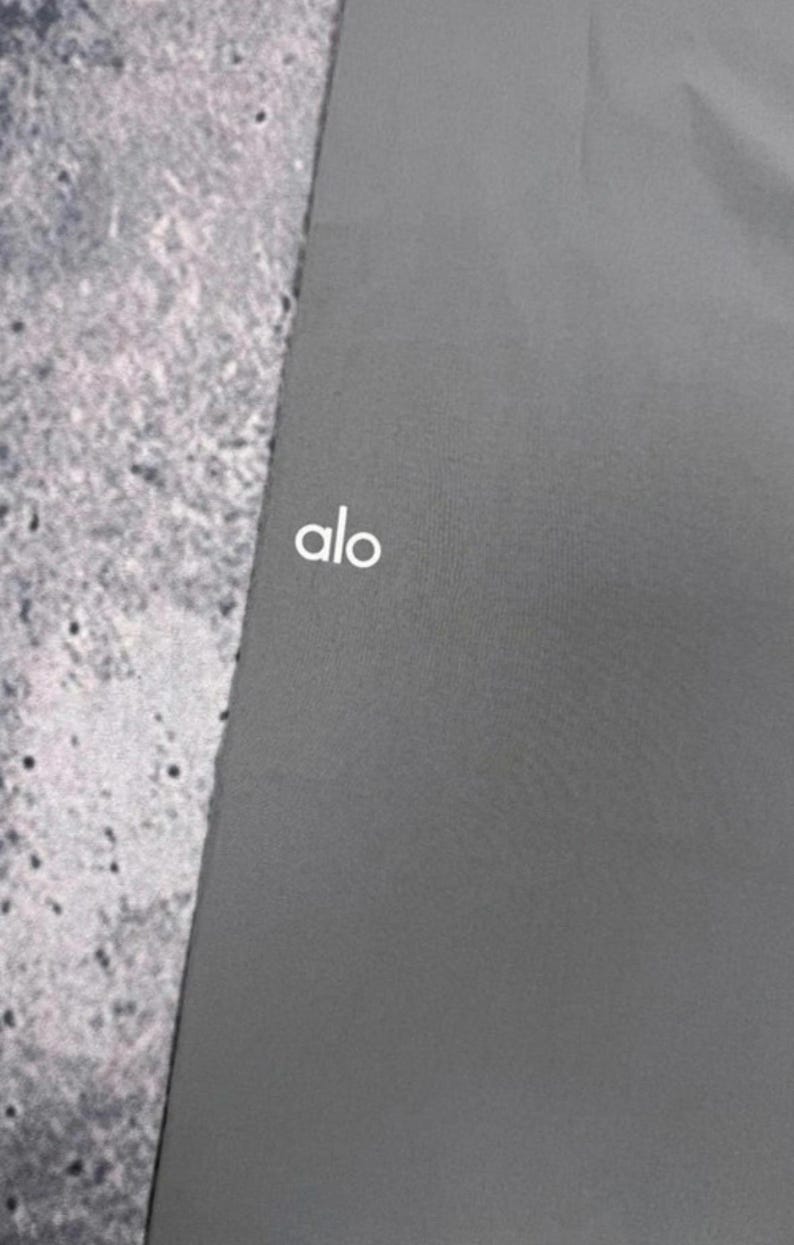 Puede incluir: Una esterilla de yoga gris oscuro con el texto blanco "alo" en la esquina superior izquierda. La esterilla es de color liso y parece estar hecha de un material liso. El fondo es una superficie texturizada de color gris claro.