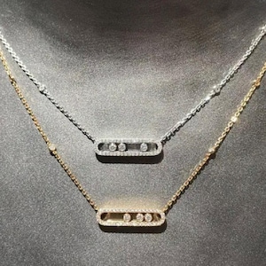 Puede incluir: Dos delicados collares con colgantes rectangulares. Uno es plateado, el otro dorado. Cada colgante presenta tres piedras transparentes dispuestas en fila. Los collares se exhiben sobre una superficie gris oscuro.