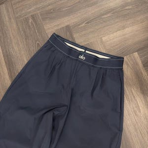 Puede incluir: Pantalones deportivos azul marino oscuro con una cintura blanca con el nombre de la marca "alo". Los pantalones tienen un corte holgado y est&aacute;n hechos de un material liso. Los pantalones est&aacute;n doblados sobre una superficie de madera.