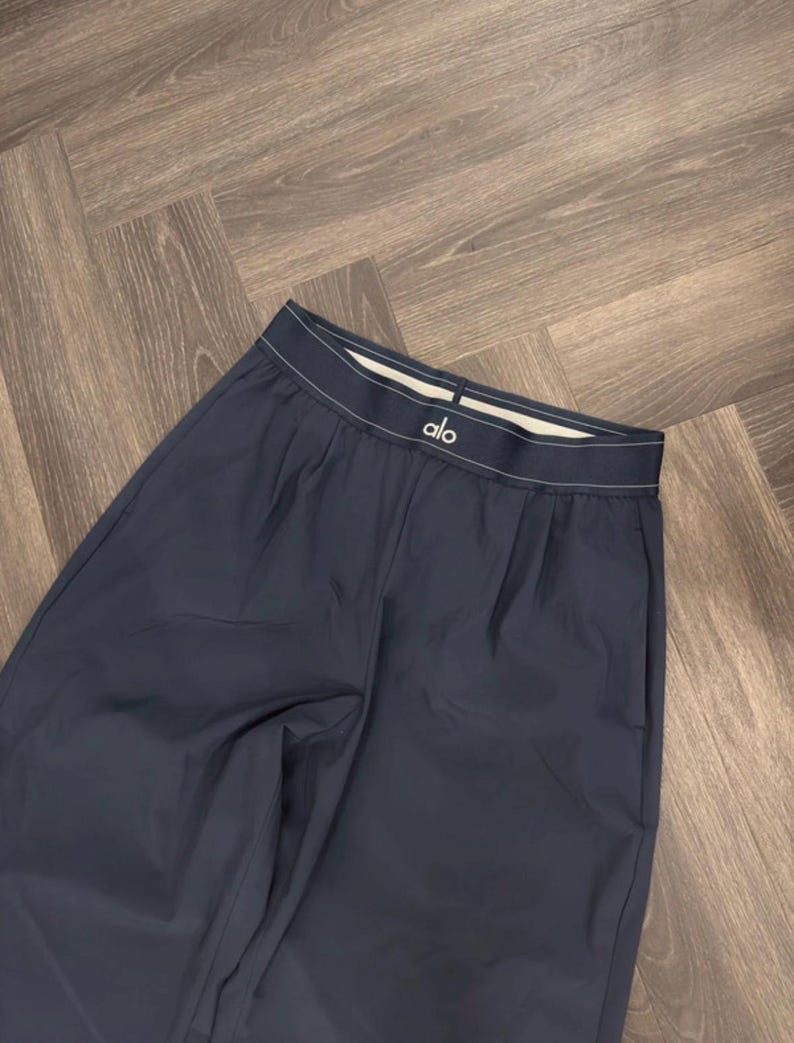 Puede incluir: Pantalones deportivos azul marino oscuro con una cintura blanca con el nombre de la marca "alo". Los pantalones tienen un corte holgado y est&aacute;n hechos de un material liso. Los pantalones est&aacute;n doblados sobre una superficie de madera.