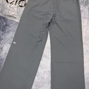 Puede incluir: Pantalones deportivos grises de pierna ancha con cintura con cord&oacute;n. Los pantalones tienen un logotipo blanco en la pierna izquierda. El nombre de la marca "alo" es visible en el embalaje.