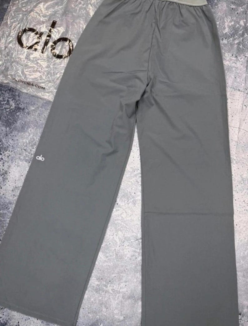 Puede incluir: Pantalones deportivos grises de pierna ancha con cintura con cord&oacute;n. Los pantalones tienen un logotipo blanco en la pierna izquierda. El nombre de la marca "alo" es visible en el embalaje.