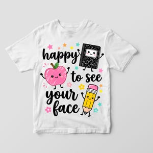 May include: A white t-shirt featuring a graphic print with the text "happy to see your face" in black script. The design includes a pink apple, a black composition notebook, and a yellow pencil, all with smiling faces, surrounded by colorful stars.