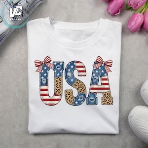 Puede incluir: Una camiseta blanca con las letras USA en un estampado gráfico grande. Las letras están decoradas con una mezcla de rayas de la bandera estadounidense, estrellas blancas sobre fondo azul y estampados de leopardo, con lazos a rayas rojas y blancas.