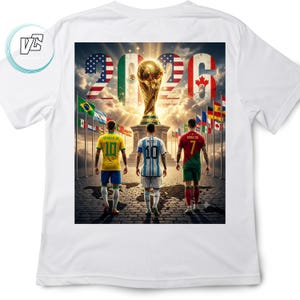 Op de afbeelding: Een wit t-shirt met een grafische print van drie voetballers voor een gouden trofee. De achtergrond toont het jaar 2026 met de vlaggen van de VS, Mexico en Canada, samen met diverse internationale vlaggen en een wereldkaart.
