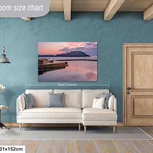 Op de afbeelding: Een ingelijste landschapsprint met een kustlandschap bij zonsondergang, met een kasteel en waterreflectie. De print is 101x152cm en hangt boven een beige hoekbank in een kamer met turquoise muren en een houten deur.