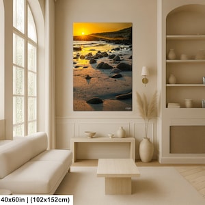 May include: A rectangular wall art print featuring a sunset over a rocky beach with golden light reflecting on the sand and water. The artwork measures 40 by 60 inches and displays a bright yellow sun low on the horizon above dark stones.