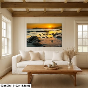 May include: A rectangular wall art print featuring a vibrant sunset over a rocky beach with golden light reflecting on the wet sand and ocean waves. The landscape photograph measures 40 inches by 60 inches and is displayed above a white sofa.