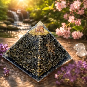 May include: A clear resin pyramid containing black stone fragments and gold flakes. The apex features a copper coil, and a gold-coloured Metatron's Cube symbol is embedded on one side. The pyramid has a smooth, polished finish and a square base.