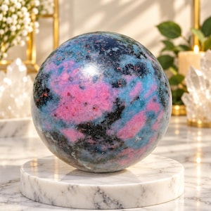 Può includere: Una pietra sferica lucida caratterizzata da un mix vibrante di inclusioni minerali rosa brillante, blu cielo e nero intenso. La superficie è liscia e riflettente, mostrando un motivo marmorizzato naturale con distinte transizioni di colore.