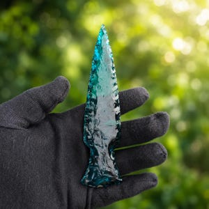 May include: A translucent teal arrowhead with a sharp, pointed tip and serrated edges. The surface of the arrowhead displays a smooth, glass-like texture with visible conchoidal fractures, creating a reflective and glossy appearance throughout.