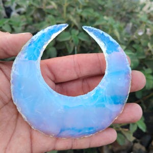 Pode incluir: Um cristal opalescente em forma de crescente. A pedra preciosa é azul pálido com áreas de azul mais escuro e branco, assemelhando-se a uma lua. O cristal é segurado em uma mão contra um fundo de folhagem verde.