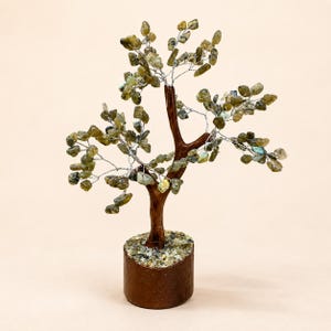 Könnte beinhalten: Eine dekorative Baumskulptur mit einem braunen Stamm und silbernen Drahtzweigen, die mit kleinen, getrommelten grünen und grauen Edelsteinen besetzt sind. Der Baum hat einen zylindrischen braunen Sockel, der mit passenden Steinsplittern bedeckt ist.