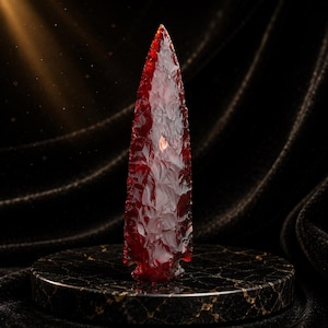 May include: A translucent, deep red arrowhead with a sharp, pointed tip and textured, flaked edges. The object is positioned vertically, displaying a glossy finish that catches light, revealing variations in the rich crimson color throughout.
