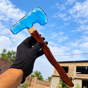 May include: A hand axe featuring a translucent, vibrant blue glass blade with a jagged, knapped edge. The blade is securely attached to a smooth, polished dark brown wooden handle with light-colored twine wrapped around the connection point.