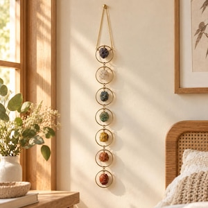 May include: A vertical wall hanging featuring seven gold-toned metal rings connected by small links. Each ring contains a single raw, rough-cut gemstone in varying colours including purple, white, blue, green, yellow, orange, and red.
