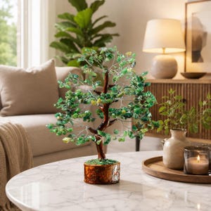 May include: A decorative tree sculpture featuring a brown trunk and silver wire branches adorned with numerous small, polished green gemstone chips. The tree is mounted on a cylindrical base with a textured, bark-like brown finish.