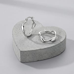 May include: Two silver hoop earrings with a fluid, irregular shape. The earrings are presented on a grey, heart-shaped concrete block. The earrings have a shiny finish and are set against a pale grey backdrop. Each earring has a clasp.