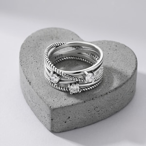 May include: A silver ring featuring three clear gemstones and a braided rope design. The ring comprises multiple intersecting bands, creating a layered effect. The ring is marked with "S925". It rests on a grey, heart-shaped surface.