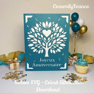 May include: A teal rectangular card featuring a white die-cut tree design with a heart at the center and the text Joyeux Anniversaire printed in white. The card is surrounded by gold and silver star confetti, a small gift box, and a cupcake.