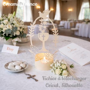 Peut inclure: Un photophore crème avec un motif découpé au laser représentant un calice, une croix et une colombe. Le photophore est sur une nappe blanche, avec un petit bouquet de fleurs blanches et des amandes enrobées.