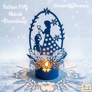 Può includere: Un portacandele blu ritagliato in carta con la silhouette di una donna e un pupazzo di neve, con un fiore a forma di cuore. Il design include fiocchi di neve e accenti floreali. Il testo "Fichier SVG Cricut Download" e "Creamily France" sono visibili.