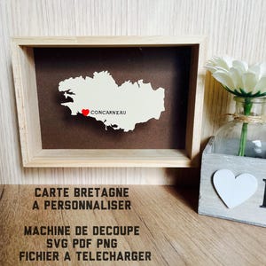Peut inclure: Une carte encadrée de la Bretagne, France, avec un cœur marquant Concarneau. La carte est blanche sur un fond marron, dans un cadre en bois clair. Le texte en bas indique "CARTE BRETAGNE A PERSONNALISER".