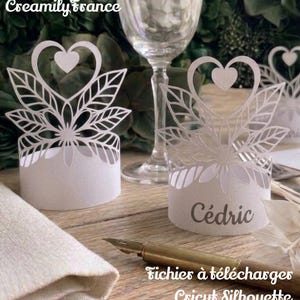 Può includere: Porta segnaposto in carta bianca con un design a cuore e foglie. Un porta segnaposto ha il nome "Cédric" stampato sopra. L'immagine include anche un bicchiere, una penna e il testo "Fichiers à télécharger Cricut Silhouette".