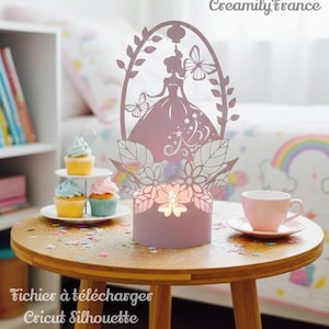 Könnte beinhalten: Eine rosa dekorative Lampe im Prinzessinnen-Stil mit einem Silhouette-Design, das eine Prinzessin, Schmetterlinge und florale Akzente zeigt. Die Lampe steht auf einem kleinen Holztisch, mit Cupcakes und einer Teetasse in der Nähe. Der Text "Creamily France" und "Fichiers à télécharger Cricut Silhouette" sind sichtbar.