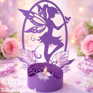 Puede incluir: Un portavelas de silueta de hada de papel morado con una vela encendida en su interior. El hada sostiene una mariposa. La base tiene recortes florales. Las palabras "Fichier SVG Téléchargement Cricut" están en la parte inferior.