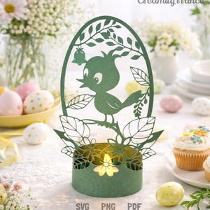 Op de afbeelding: Een groene papieren paasei-vormige kandelaar met een uitgesneden ontwerp van een vogel die op een tak zit. De kandelaar heeft een bloemenbasis en de woorden "SVG PNG PDF" onderaan. Paaseieren en cupcakes staan op de achtergrond.