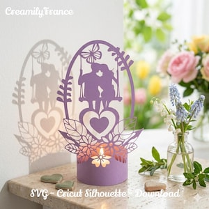 Puede incluir: Un portavelas de papel lavanda con la silueta de una pareja en un columpio, un corazón y detalles florales. El portavelas proyecta una sombra en la pared. Hay una vela encendida dentro. Las palabras "Creamily France" y "SVG - Cricut Silhouette - Download" son visibles.