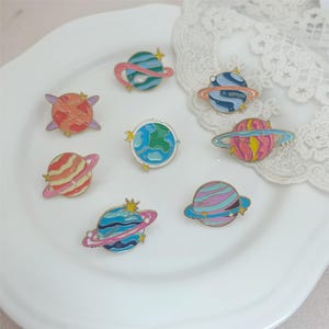 May include: A selection of colourful enamel pins depicting planets and celestial bodies. The pins feature diverse designs, including planets with rings, stars, and the Earth. The pins are arranged on a white plate, with a lace doily in the background.