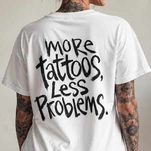 Peut inclure: Un t-shirt blanc à manches courtes avec un texte noir écrit à la main au dos qui dit "more tattoos, Less Problems." Le t-shirt a une encolure ronde et une coupe décontractée, affichant une typographie audacieuse de style pinceau au dos.