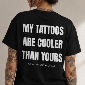 Peut inclure: Un t-shirt noir à manches courtes avec le texte blanc "MY TATTOOS ARE COOLER THAN YOURS" en police sans-serif grasse, suivi du texte plus petit en écriture cursive "but we can still be friends" imprimé au dos du vêtement.