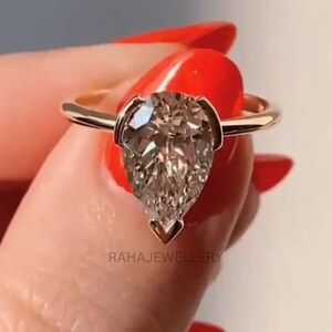 Könnte beinhalten: Ein goldener Ring mit einem großen, birnenförmigen Diamanten. Der Diamant ist in einer zarten Fassung gefasst. Der Ring wird vor einem neutralen Hintergrund gezeigt.