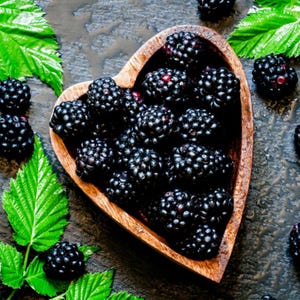 May include: A heart-shaped wooden bowl filled with fresh, ripe blackberries. The dark, juicy berries are surrounded by green leaves, creating a natural and appealing arrangement. The berries are a deep, rich black color.