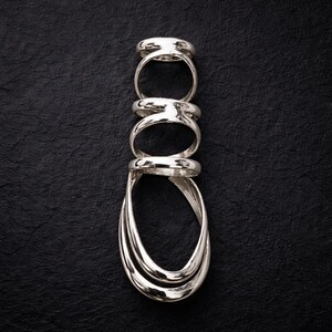 May include: A stack of five silver rings arranged vertically against a black textured background. The rings have a polished, reflective surface, with varying designs including a rounded band and a layered, elongated oval shape.