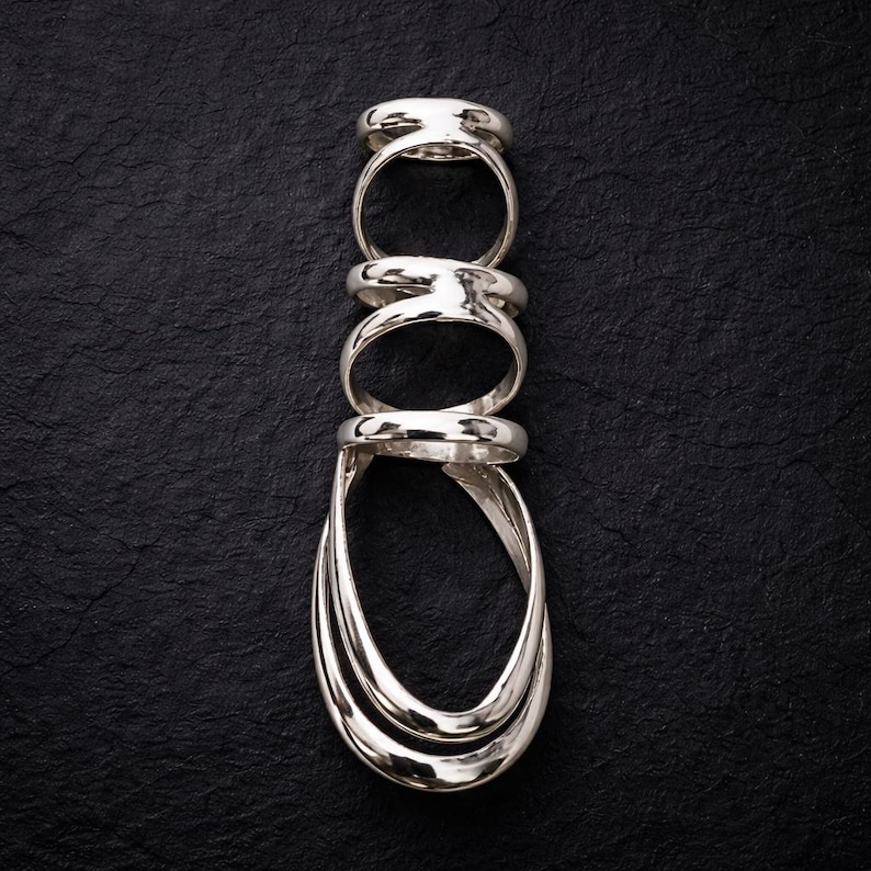May include: A stack of five silver rings arranged vertically against a black textured background. The rings have a polished, reflective surface, with varying designs including a rounded band and a layered, elongated oval shape.