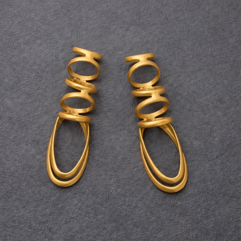 May include: A pair of gold-colored earrings. Each earring features a series of connected rings leading to a double teardrop shape. The earrings are displayed against a dark gray background.