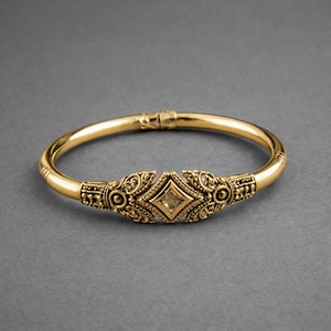 May include: A gold-coloured bangle bracelet with intricate detailing. The bracelet has a central diamond-shaped design with a clear stone and decorative patterns. The clasp is visible at the top of the bracelet.